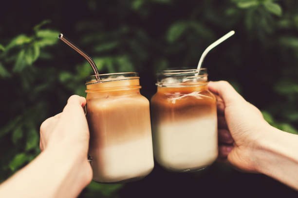 Two glass jars of ice coffee metal straws in woman and man hands. Clink cups of coffee. Outdoor shot. Lifestyle, picnic vibes. Trendy toning. Copy space for your text.