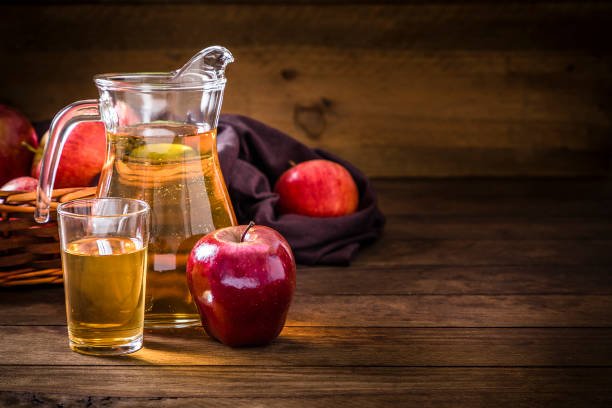 Front view of a crystal jug and a drinking glass full of apple juice on a rustic wooden table. In front of the pitcher is a single red apple and behind is a wicker basket full of multicolored apples. The objects are at the left side of the image leaving a useful copy space at the right on the table. Low key DSLR photo taken with Canon EOS 6D Mark II and Canon EF 24-105mm f / 4