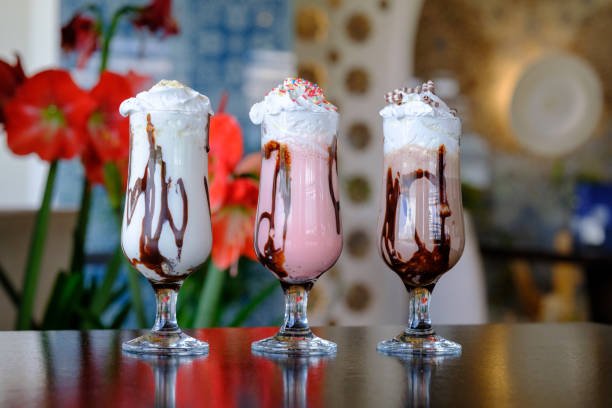 Three milkshakes with whipped cream and chocolate drizzle, displayed on a dark surface. They are different flavors: vanilla, strawberry, and chocolate.